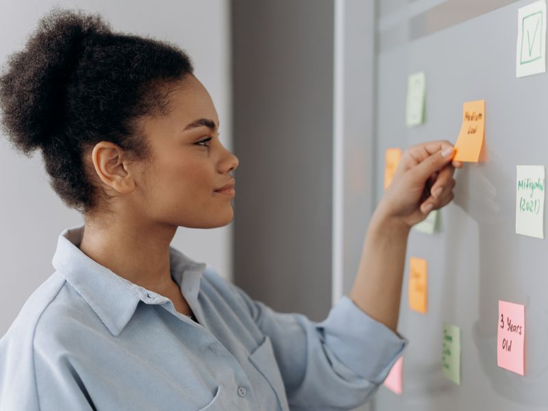 A woman in a light blue shirt, seen in profile, placing an orange sticky note onto a grey wall covered with pa