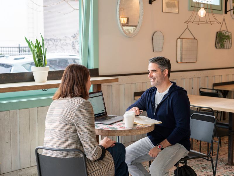 A woman in a checked blazer, seen from behind, sits across a small round cafe table from a smiling man in a na