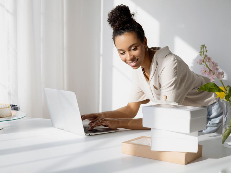 A woman leans over a white table in a bright, sunlit room, smiling as she types on a laptop with one hand whil