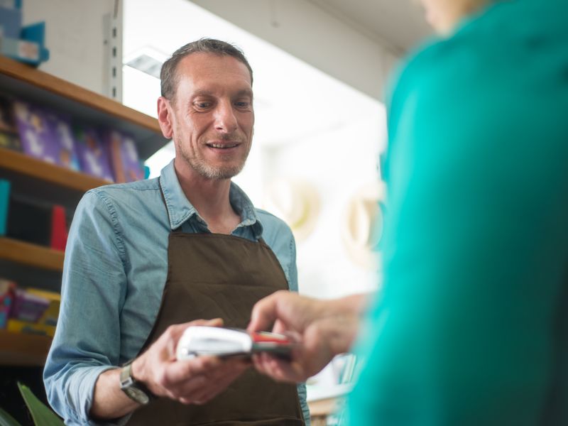 A smiling shop worker in a denim shirt and brown apron holds out a small white card payment terminal toward a 